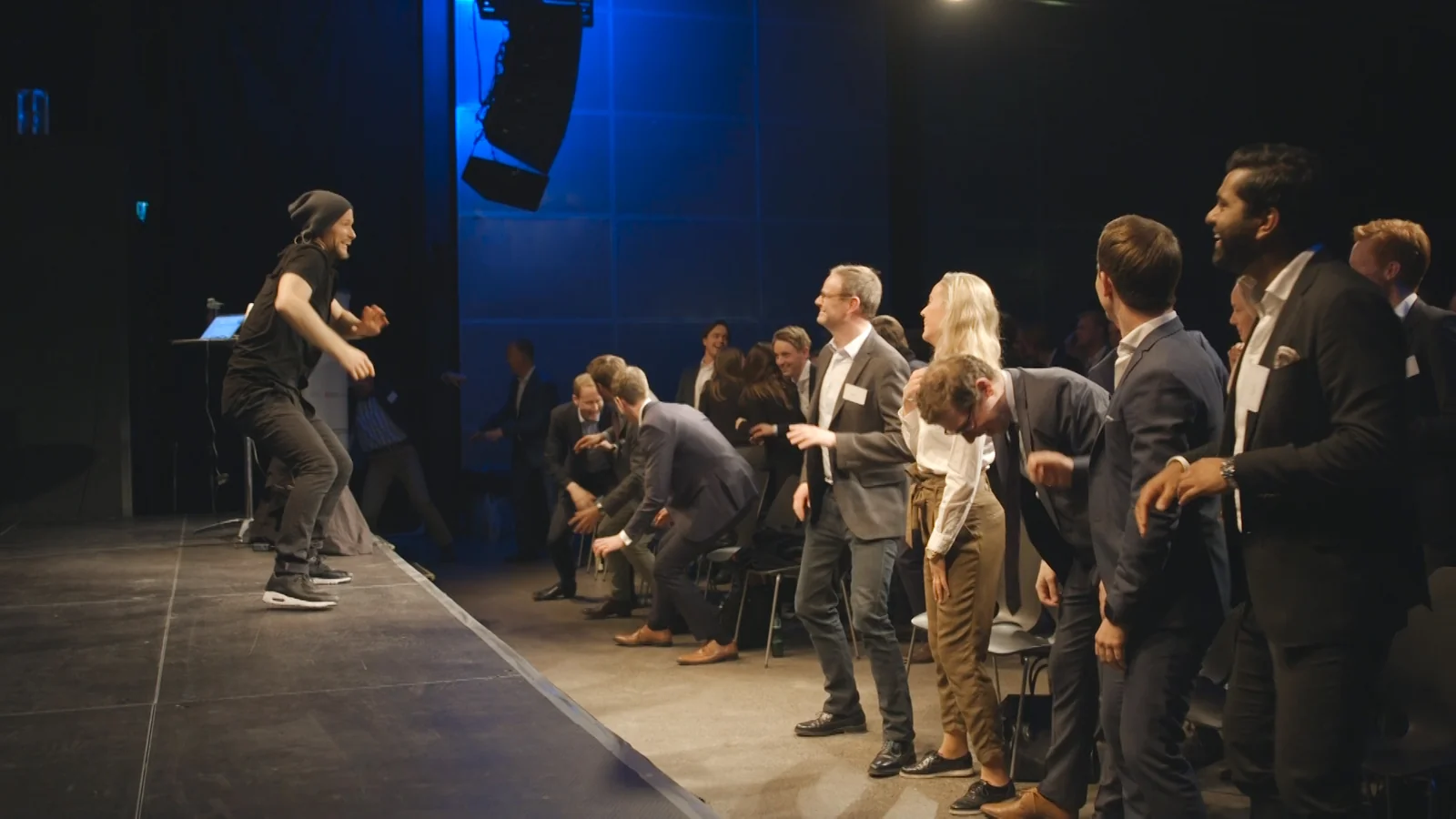 Kyrre Texnæs dancing on stage while executives in suits stand up and move with him