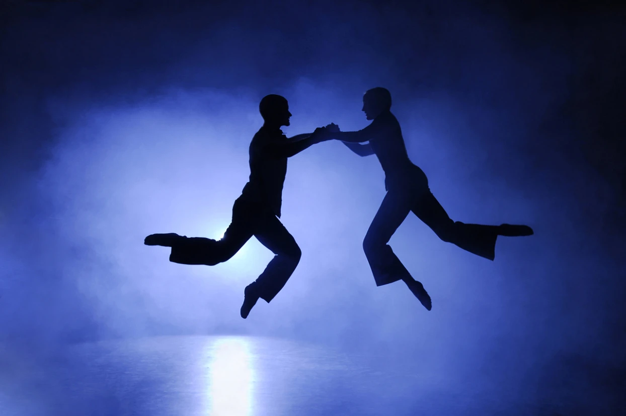 Two dancers meet hand in hand mid-jump, silhouetted against light
